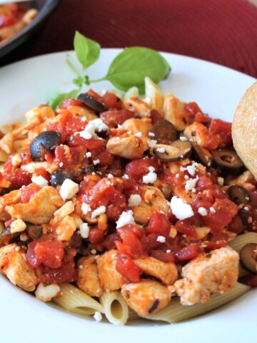 Pasta, red sauce, black olives, and chicken in a white bowl, with basil and a soft roll.