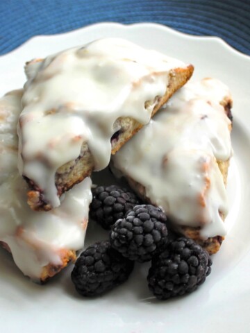 Three blueberry scones with glaze on a white plate, along with some fresh blackberries.