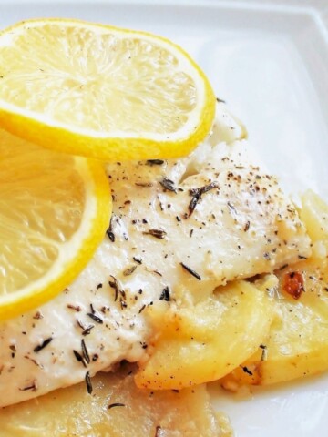 Fresh fish with herbs on top of roasted potatoes, with lemon slices on top.