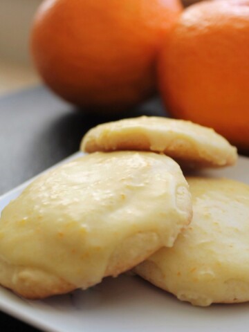 Three orange cookies with orange frosting and cara cara oranges in the background.