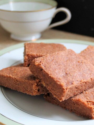 Chocolate shortbread stacked on white plate with a tea cup in the background.
