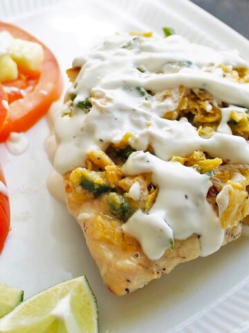 Baked chicken with a crunchy topping and lime sour cream sauce on a white plate with sliced fresh tomatoes.