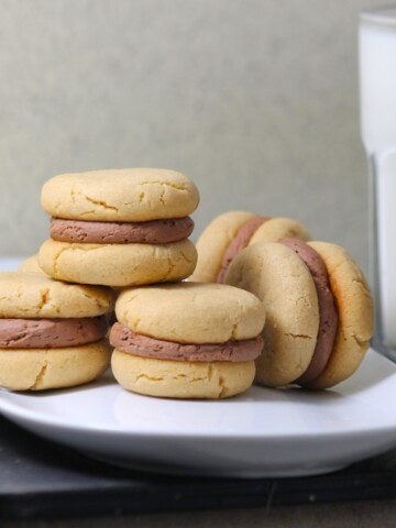 Small stack of mini peanut butter and ganache sandwich cookies with a cup of milk.