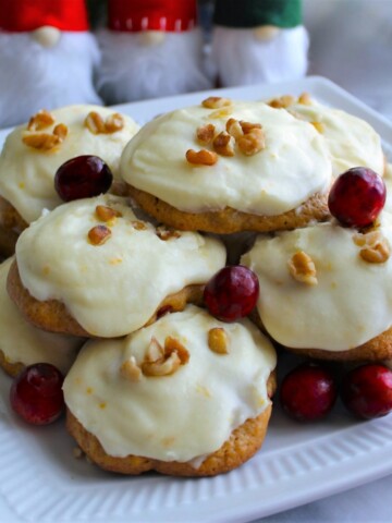 Pile of cranberry orange nut cookies on a white plate with 3 tiny Santas in the background.