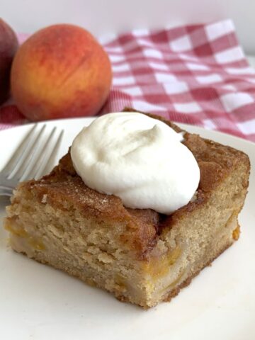 Slice of Fresh Peach cake with a dollop of whipped cream. On a small white plate with a couple of peaches in the background.